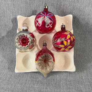 May include: Four vintage Christmas ornaments in a white egg crate. The ornaments are red, silver, and gold, with designs including bells, flowers, and a starburst. A festive holiday decoration.