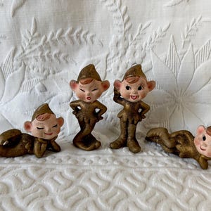 May include: Four vintage ceramic elf figurines in various poses. The elves have rosy cheeks, brown hair, and are dressed in gold-colored outfits. The figurines are displayed on a white, textured surface.