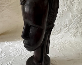 May include: A dark brown wooden sculpture of a stylized human head. The sculpture features detailed carving of the hair and facial features, with a smooth, polished finish. The head rests on a cylindrical base, showcasing a classic African art style.