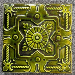 May include: A square ceramic tile featuring a glossy olive green glaze with a raised relief pattern. The design includes a central circular floral motif surrounded by geometric borders, rope-like edges, and stylized corner accents with deep shadows.