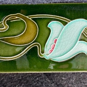 May include: A rectangular ceramic tile with a glossy dark green glaze and a crackle finish. The tile features a raised relief design of a light blue flower with pink accents and brown stems, with the letter A printed in the bottom right corner.