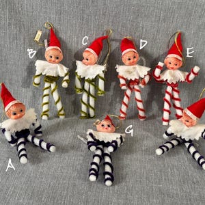 May include: A collection of vintage Christmas elf ornaments. Each elf has a red hat, a white ruffled collar, and a painted face. The elf's bodies are made of chenille stems in various colors, including green, red, and blue.
