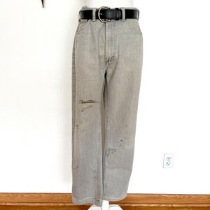 Pode incluir: Cal&ccedil;a de ganga cinzenta com cinto de couro preto. A cal&ccedil;a tem um visual desgastado com v&aacute;rias manchas castanhas claras.