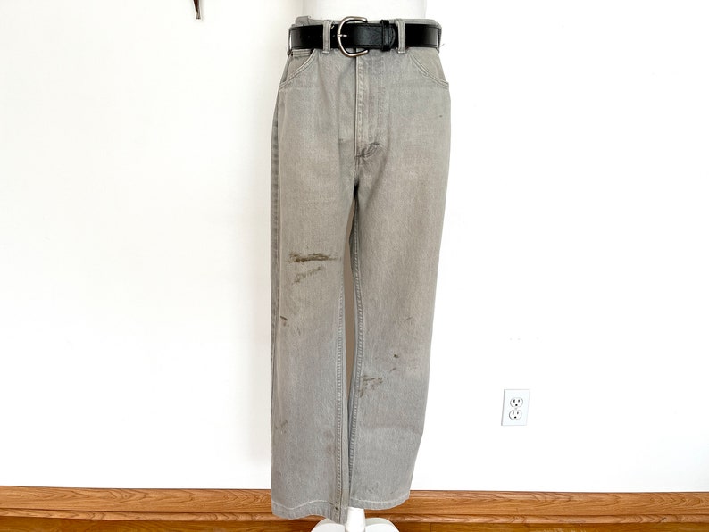 Pode incluir: Cal&ccedil;a de ganga cinzenta com cinto de couro preto. A cal&ccedil;a tem um visual desgastado com v&aacute;rias manchas castanhas claras.