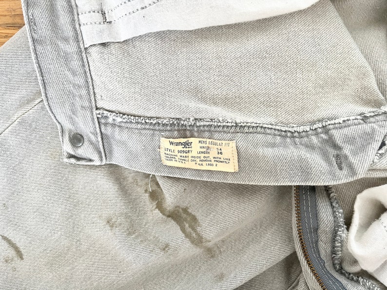 Pode incluir: Um par de cal&ccedil;as de ganga Wrangler cinzentas claras com uma mancha castanha. As cal&ccedil;as de ganga t&ecirc;m um fecho de correr e um r&oacute;tulo que diz "Wrangler Men's Regular Fit Style 309GRY Waist 34 Length 36 Machine Wash Inside Out With Like Colors Tumble Dry Remove Promptly Made In U.S.A. L.N. 2"