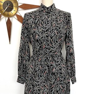 May include: A long-sleeved button-down dress featuring a black base with a repeating pattern of thin, angular leaf-like shapes in white, light blue, and soft pink. The dress has a structured collar, long sleeves with cuffs, and a matching fabric belt.