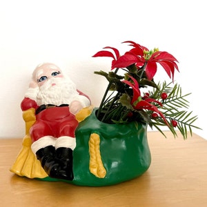 May include: A ceramic planter shaped like Santa Claus's sack, with a Santa Claus figurine sitting on top. The planter is green with yellow trim and is filled with a red poinsettia plant and green foliage.
