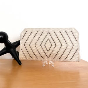 May include: A rectangular off-white clutch bag featuring a textured beaded surface with a geometric diamond pattern created by dark grey beads. The bag has a structured shape with angled top corners and is displayed on a clear plastic stand.
