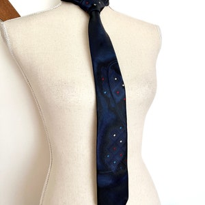 May include: A dark navy blue necktie featuring a subtle, darker abstract pattern and small, scattered square accents in red, white, and light blue. The tie is tied in a knot and displayed on a cream-colored mannequin torso against a plain background.