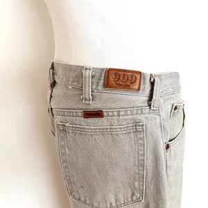 Pode incluir: Jeans Wrangler 909 cinzento com um remendo de couro marrom que diz "909 Wrangler".