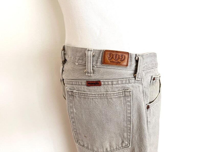 Pode incluir: Jeans Wrangler 909 cinzento com um remendo de couro marrom que diz "909 Wrangler".
