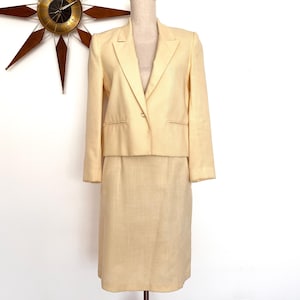 May include: A pale yellow skirt suit, comprising a blazer and a knee-length skirt. The blazer features a single-button closure and a notched lapel. The suit is displayed on a mannequin against a white backdrop. A mid-century modern clock is visible in the background.
