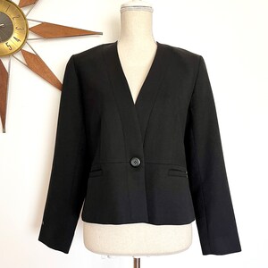 May include: A solid black blazer featuring a deep V-neckline, long sleeves, and a single central button closure. The garment has a structured design with horizontal seam detailing at the waist and two small horizontal slit pockets on the front.