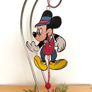 May include: A flat, two-dimensional hanging ornament featuring a cartoon mouse character wearing a black top hat with a blue and red band, a red vest, blue pants, and large yellow shoes. The character is suspended by a thin red string.