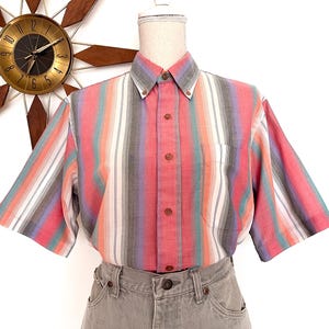 May include: A short-sleeved shirt with vertical stripes in pink, red, grey, white, and teal. The shirt has a button-down collar and a single chest pocket. The shirt is displayed on a mannequin.