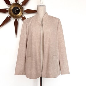 Puede incluir: Blazer beige claro con frente abierto, cuello mandarín y dos bolsillos delanteros. La chaqueta se exhibe en un maniquí sobre un fondo blanco. Un reloj de estrella de mediados de siglo cuelga en la pared detrás del maniquí.