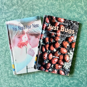 May include: Two children's books with colorful covers. The book on the left is titled "You Need Your Nose" and features a photo of two children smelling a flower. The book on the right is titled "Just Bugs" and features a photo of ladybugs.