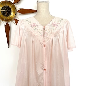 May include: A light pink vintage nightgown with short sleeves and a button-down front. The neckline is adorned with embroidered floral details. The nightgown is displayed on a mannequin, with a mid-century modern clock in the background.