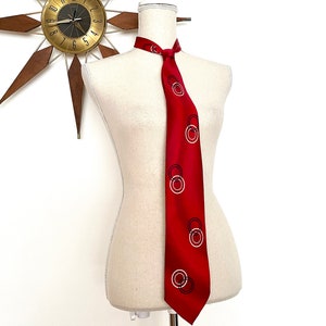 May include: A vibrant red necktie featuring a repeating pattern of interlocking white and black circular rings. The tie is displayed on a neutral-toned mannequin torso, showcasing the smooth fabric texture and the geometric design printed across the length.
