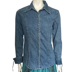 May include: A light blue denim shirt with long sleeves and a pointed collar. The shirt features a button-down front and decorative stitching along the seams. The cuffs have tie details. The shirt is a classic style.