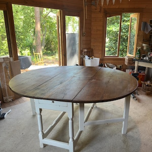 May include: A large round drop-leaf table featuring a dark brown stained wooden top with a visible grain pattern. The table is supported by a sturdy white painted base with turned legs and a small integrated drawer on one side of the frame.