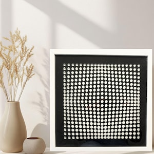 Könnte beinhalten: Ein quadratisches Wandkunstwerk mit einem weißen Rahmen, der eine schwarze Platte mit einem Raster aus gleichmäßig verteilten weißen kreisförmigen Punkten umschließt. Die Punkte sind in einem präzisen Muster angeordnet.
