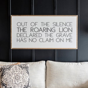 Może przedstawiać: Biała kanapa z tkaniny z drewnianym stelażem i dwoma poduszkami. Kanapa stoi przed czarną ścianą z ramą z napisem "Out of the silence the roaring lion declared the grave has no claim on me."