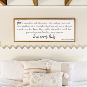 Op de afbeelding: Een wit houten bord met een bruin frame hangt boven een wit bed met een wit hoofdeinde. Het bord heeft een grijze inscriptie die luidt: "Liefde is geduldig, liefde is vriendelijk. Ze is niet jaloers, ze is niet opschepperig, ze is niet trots. Ze handelt niet onfatsoenlijk, ze zoekt niet haar eigen belang, ze is niet snel boos, ze houdt geen rekening met kwaad. Liefde verheugt zich niet over onrecht, maar verheugt zich over de waarheid. Ze beschermt altijd, ze vertrouwt altijd, ze hoopt altijd, ze volhardt altijd. Liefde faalt nooit. 1 Korinti&euml;rs 13:4-8"