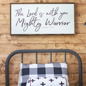 May include: A white wooden sign with black calligraphy that reads "The Lord is with you Mighty Warrior Judges 6:12" hanging on a wood paneled wall above a black metal bed frame with a black and white checkered pillow and a white pillow with black crosses.