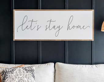 May include: A white couch with two pillows, one brown and one patterned, in front of a black wall with a wooden sign that reads "Let's stay home".