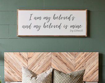May include: A bedroom with a green wall and a wooden headboard. The headboard is made of wood planks arranged in a chevron pattern. There are three pillows on the bed, two with a floral pattern and one with a striped pattern. A sign above the headboard reads "I am my beloved's and my beloved is mine Song of Solomon 6:3".