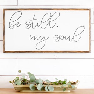May include: A white wooden sign with a brown frame, featuring the text "be still, my soul" in cursive script.