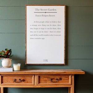 May include: A wooden console table with two drawers and a framed print with a quote from "The Secret Garden" by Frances Hodgson Burnett. The quote reads: "At first people refuse to believe that a strange new thing can be done, then they begin to hope it can be done, then they see it can be done - then it is done and all the world wonders why it was not done centuries ago." The number 118 is printed below the quote.