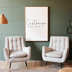 May include: Two light gray upholstered armchairs with tufted backs and gold legs, facing each other in front of a white framed sign with the text "Customize This Sign" on a green wall.