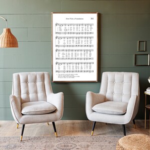 May include: Two light gray armchairs with button tufting and black metal legs sit in front of a green wall. A framed print of a hymn titled "How Firm a Foundation" hangs on the wall above the chairs.
