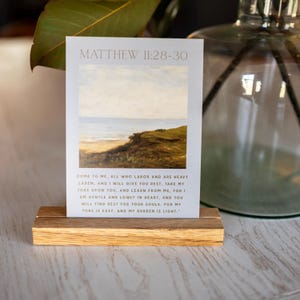 May include: A rectangular art print with a landscape painting of a beach and ocean scene, displayed on a wooden block. The print has the text "MATTHEW 11:28-30" at the top, with a Bible verse below. The wooden block is light brown.