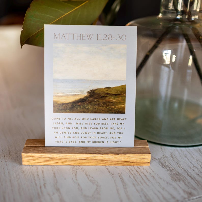 May include: A rectangular art print with a landscape painting of a beach and ocean scene, displayed on a wooden block. The print has the text "MATTHEW 11:28-30" at the top, with a Bible verse below. The wooden block is light brown.