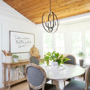 May include: A white farmhouse dining table with a round top and a white base. The table is set with a white plate and a white bowl with green leaves. There are four chairs around the table, two of which are visible in the image. The chairs are upholstered in gray fabric and have wooden frames. The table is in a room with white walls and a wooden ceiling. There is a sign on the wall that says "Taste and see that the Lord is good."
