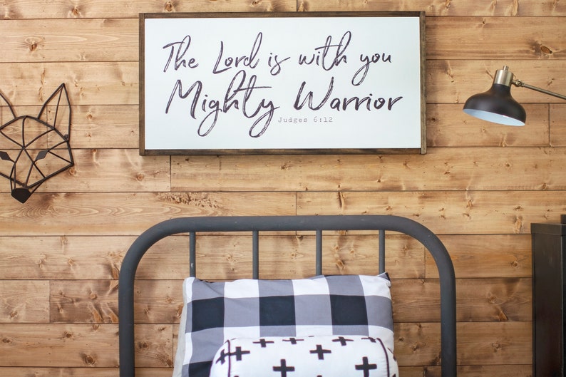 May include: A white wooden sign with black calligraphy that reads "The Lord is with you Mighty Warrior Judges 6:12" hanging on a wood paneled wall above a black metal bed frame with a black and white checkered pillow and a white pillow with black crosses.