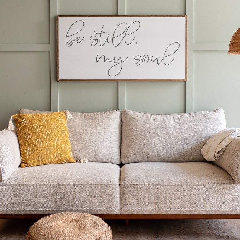 May include: A white wooden sign with the text "be still, my soul" in black cursive script. The sign is hanging on a light green wall above a light gray couch with a yellow throw pillow.