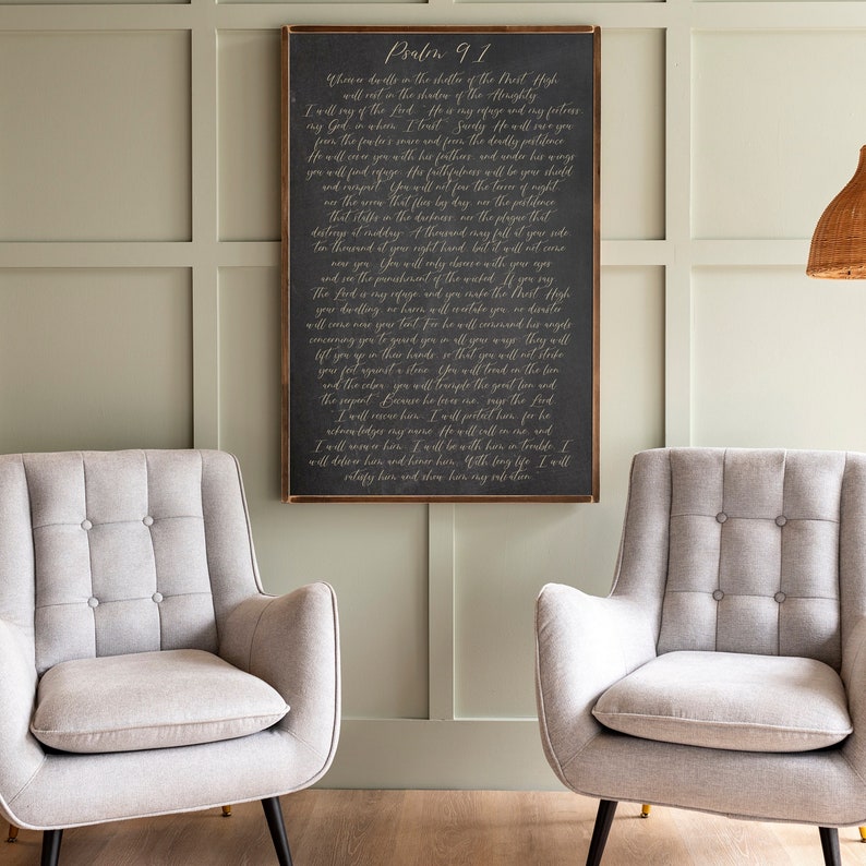 May include: A dark gray wooden sign with a white inscription of Psalm 91. The sign is hanging on a wall with a light green background. Two light gray chairs are in the foreground.