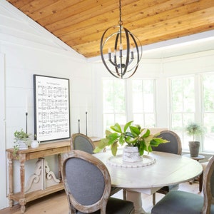 May include: A dining room with a white round table, gray upholstered chairs, and a wooden console table. A black framed print of sheet music hangs on the wall. A black orb chandelier hangs above the table. The ceiling is made of wood.