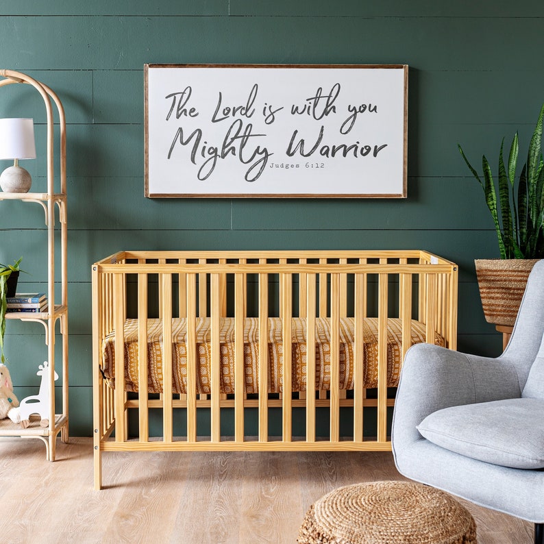 May include: A wooden crib with a mustard yellow and white patterned sheet. The crib is in front of a green wall with a framed sign that reads "The Lord is with you Mighty Warrior Judges 6:12".