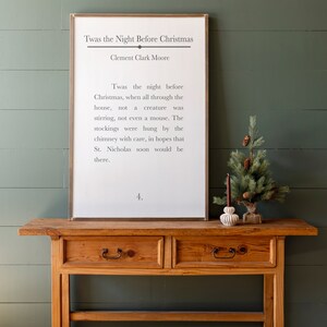 Puede incluir: Una impresi&oacute;n enmarcada blanca con el texto "Twas the Night Before Christmas" de Clement Clark Moore. El texto es un poema sobre Santa Claus y la v&iacute;spera de Navidad. La impresi&oacute;n est&aacute; sobre una mesa de madera con un peque&ntilde;o &aacute;rbol de Navidad y un portavelas.