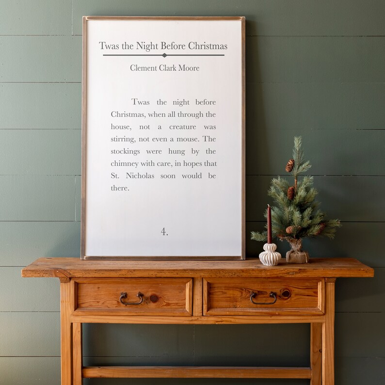 Puede incluir: Una impresi&oacute;n enmarcada blanca con el texto "Twas the Night Before Christmas" de Clement Clark Moore. El texto es un poema sobre Santa Claus y la v&iacute;spera de Navidad. La impresi&oacute;n est&aacute; sobre una mesa de madera con un peque&ntilde;o &aacute;rbol de Navidad y un portavelas.