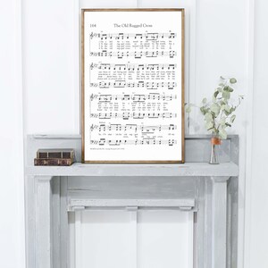 May include: A framed print of sheet music titled "The Old Ruggled Cross" with musical notes and lyrics. The frame is a warm brown color. The print is displayed on a gray mantel with a small vase of greenery.