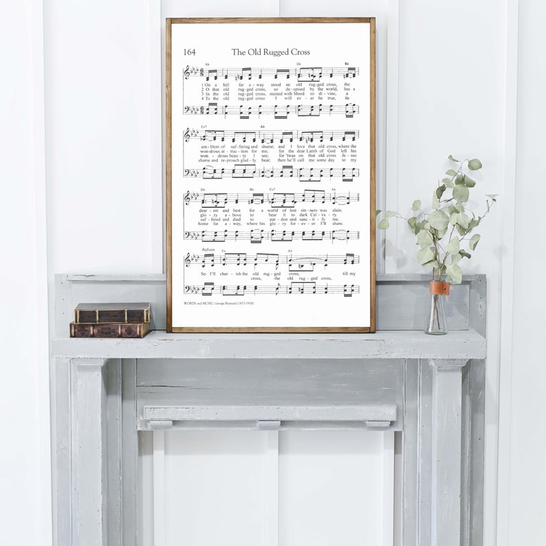May include: A framed print of sheet music titled "The Old Ruggled Cross" with musical notes and lyrics. The frame is a warm brown color. The print is displayed on a gray mantel with a small vase of greenery.
