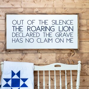 Może przedstawiać: Biały drewniany znak z napisem w kolorze granatowym, kt&oacute;ry brzmi "Out of the silence the roaring lion declared the grave has no claim on me". Znak wisi na drewnianej ścianie.