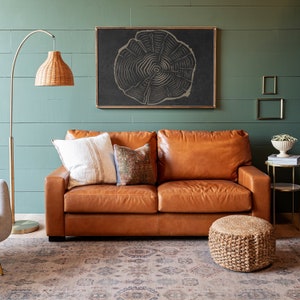 K&ouml;nnte beinhalten: Ein braunes Ledersofa mit zwei Kissen und einem Zierkissen. Das Sofa steht vor einer gr&uuml;nen Wand mit einem schwarzen gerahmten Druck eines Baumrings. Ein geflochtener Fu&szlig;hocker steht vor dem Sofa.