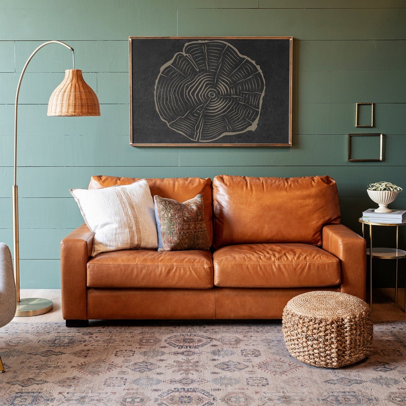 K&ouml;nnte beinhalten: Ein braunes Ledersofa mit zwei Kissen und einem Zierkissen. Das Sofa steht vor einer gr&uuml;nen Wand mit einem schwarzen gerahmten Druck eines Baumrings. Ein geflochtener Fu&szlig;hocker steht vor dem Sofa.